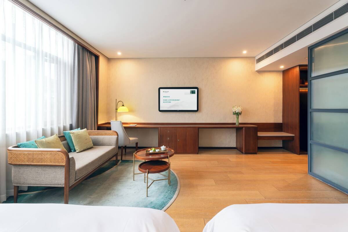 Une chambre d’hôtel avec un canapé, un bureau, une chaise, une petite table avec un vase et une grande fenêtre avec des rideaux.