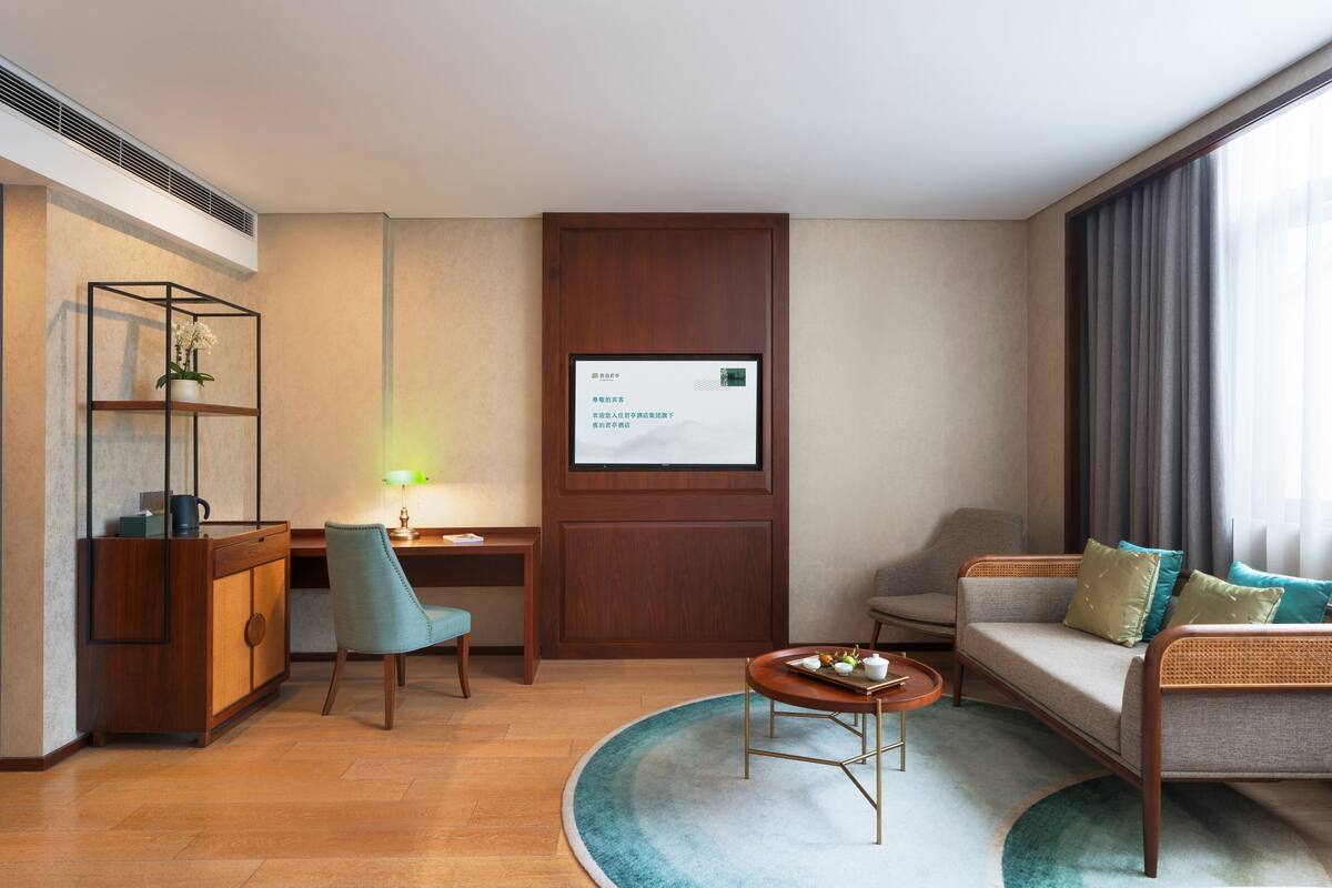 Une chambre d’hôtel moderne avec un bureau en bois, un canapé confortable, une table basse ronde et une grande fenêtre avec des rideaux.