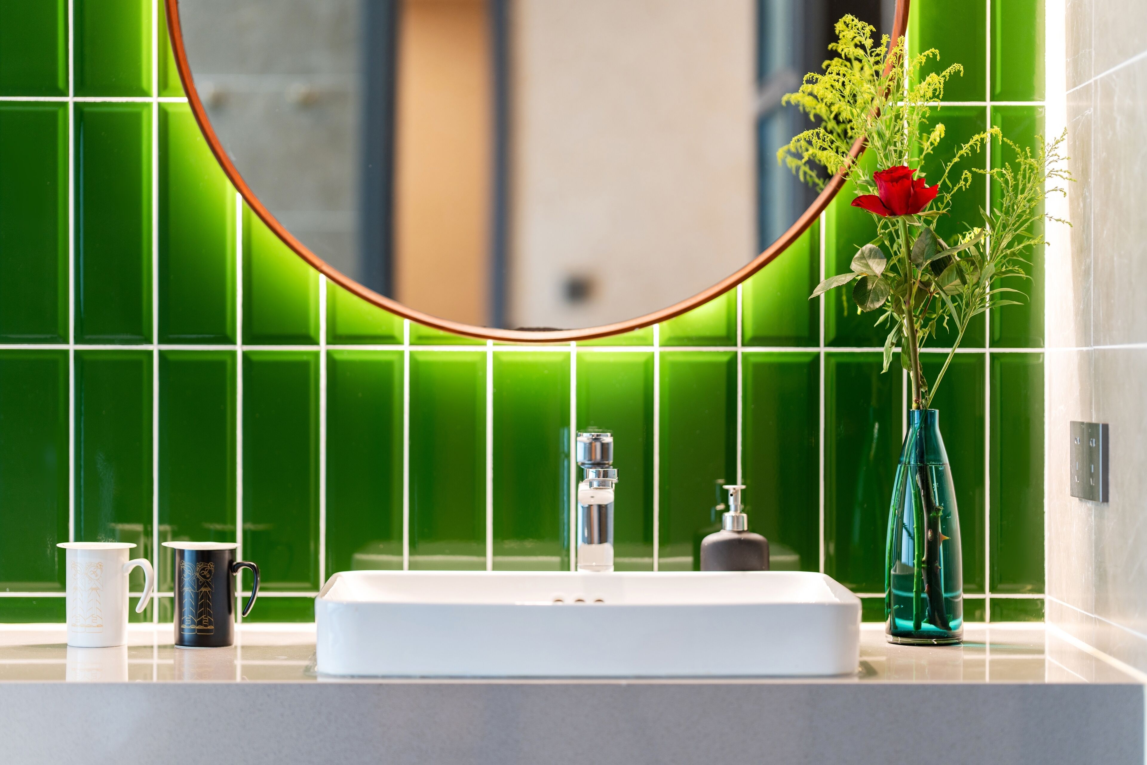 Een badkamer met een witte wasbak, groene tegels, een ronde spiegel en een vaas met bloemen.