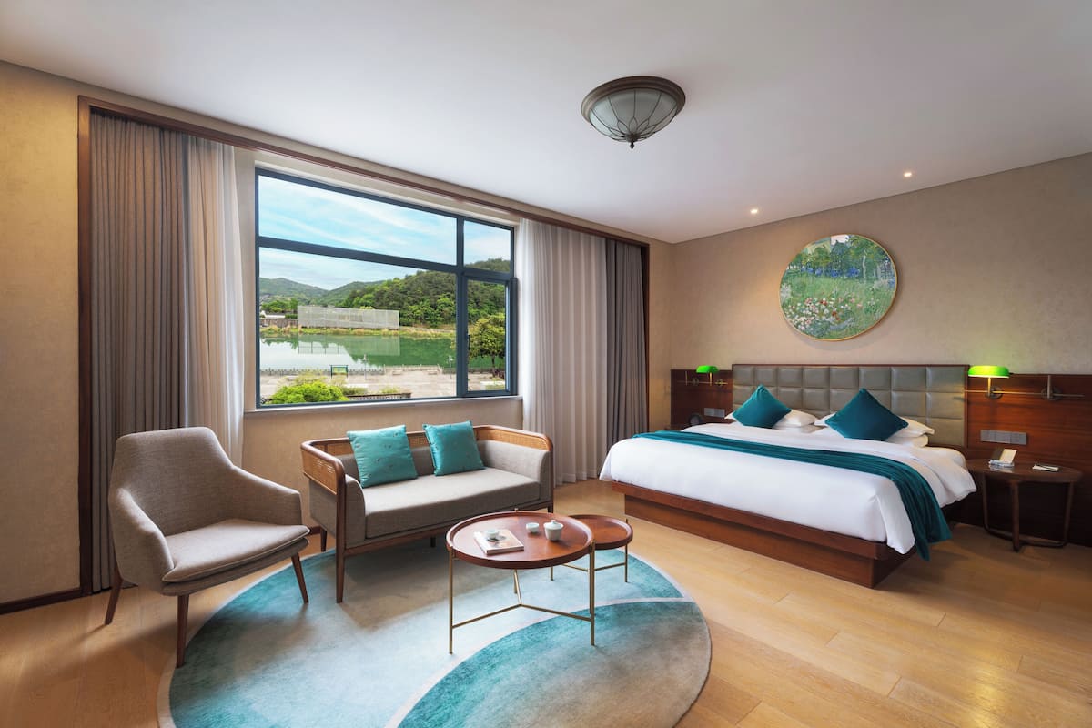 Une chambre d’hôtel avec un grand lit, un canapé, un fauteuil, une petite table et une vue sur un paysage, visible à travers une grande fenêtre.