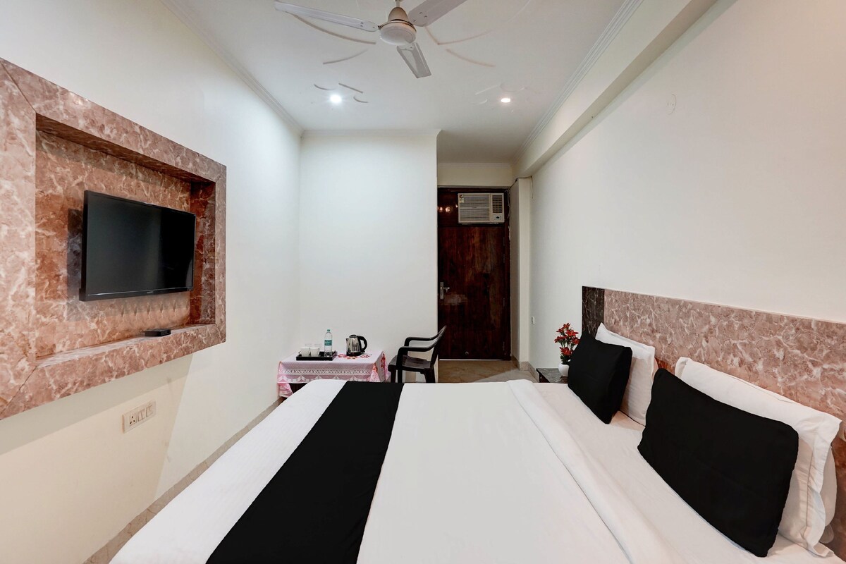A hotel room with a bed, a flat-screen TV mounted on a marble wall, a ceiling fan, a small table with a kettle, and a door with an air conditioning unit.