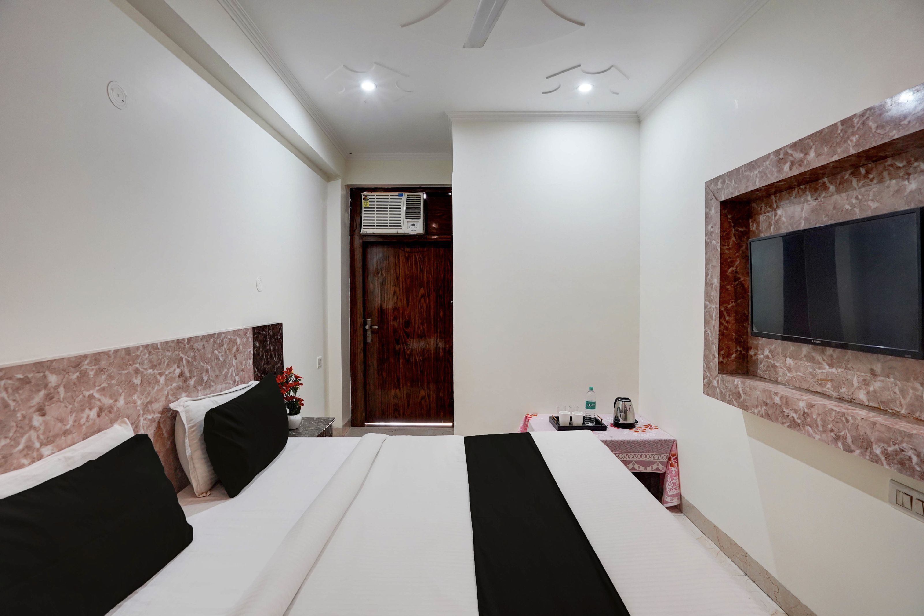 A hotel room with two beds, a TV mounted on the wall, a wooden door, and a small table with a water bottle and a kettle.