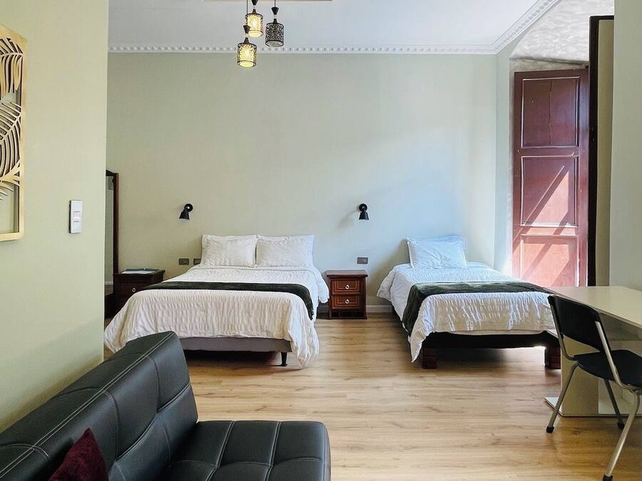 Una habitación con dos camas, un sofá negro, una mesita pequeña y un otoman colorido.