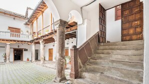 Um pátio com escadaria de pedra que leva a uma porta de madeira, rodeado por paredes brancas e vigas de madeira.