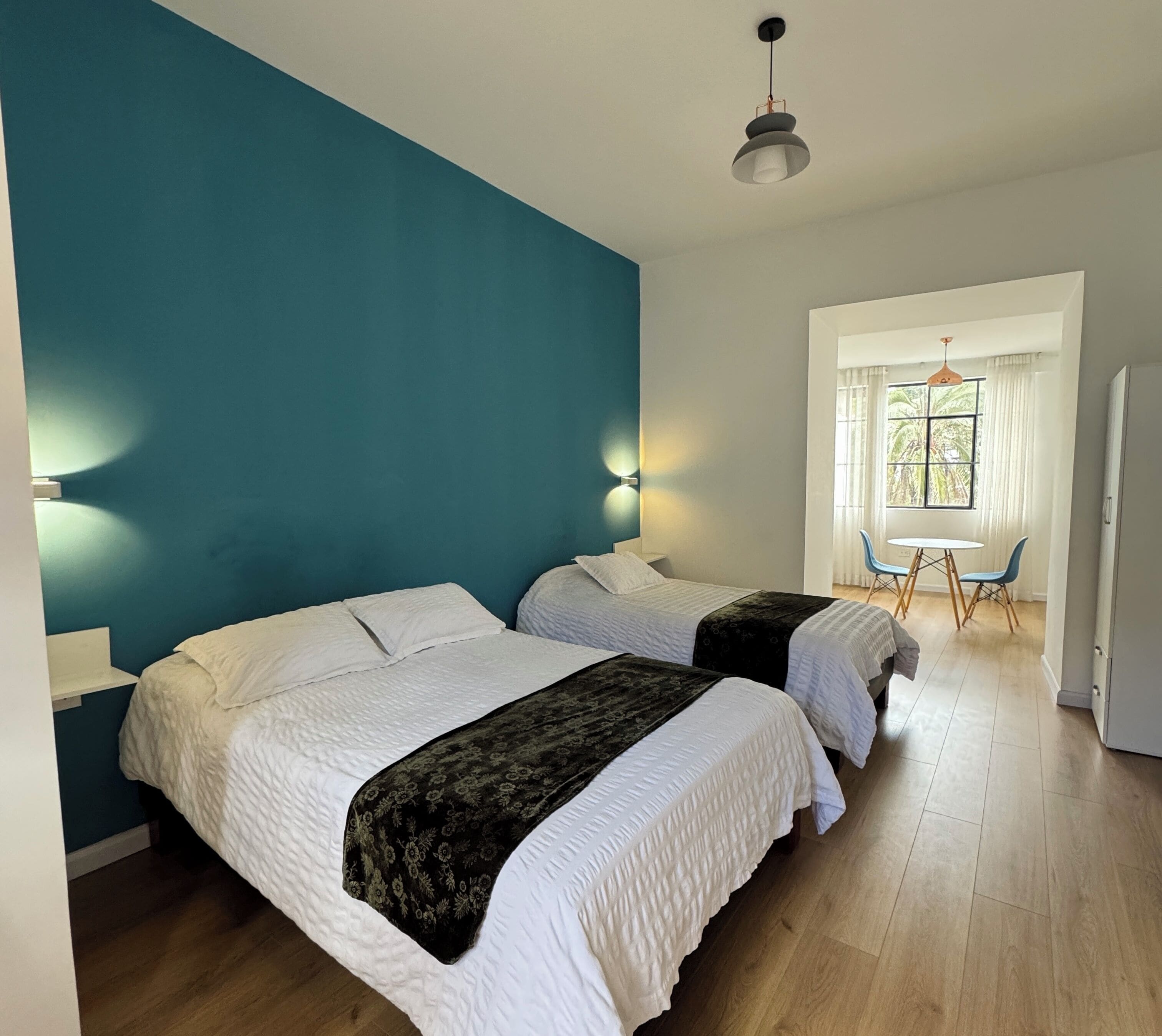 A room with two beds, a small table with chairs, a window with curtains, and a door.