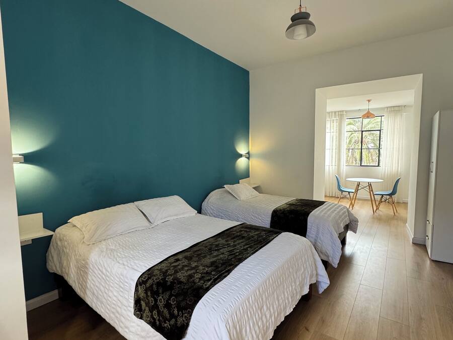 A room with two beds, a small table with chairs, a window with curtains, and a door.