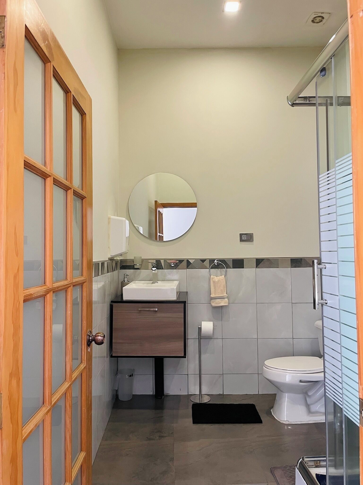 A compact bathroom with a wooden door, a round mirror, a sink, a toilet, and a shower area.