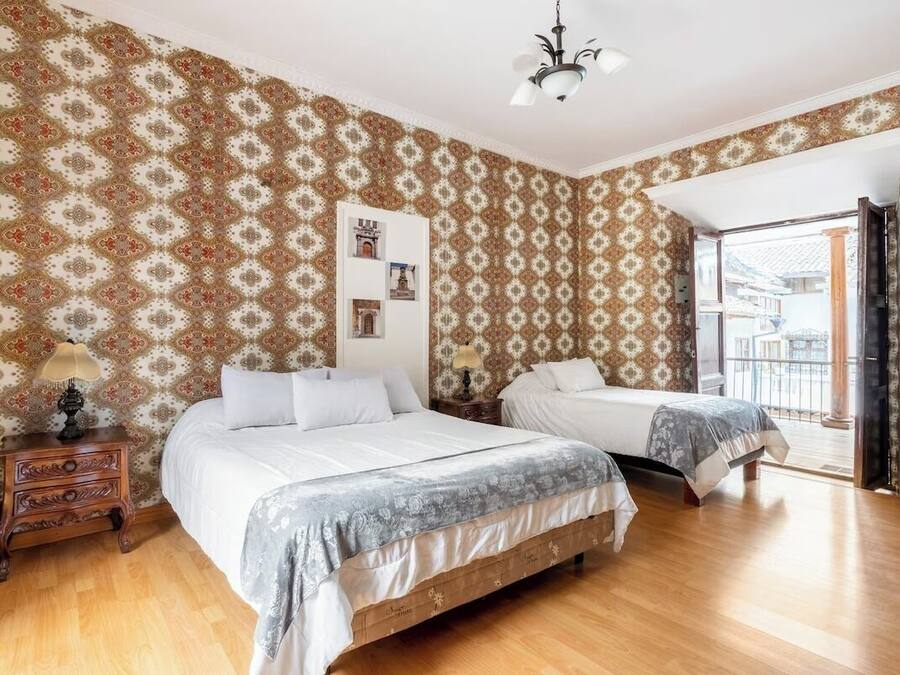 Un dormitorio con dos camas, suelo de madera, papel tapiz estampado, una araña y un balcón con vistas.