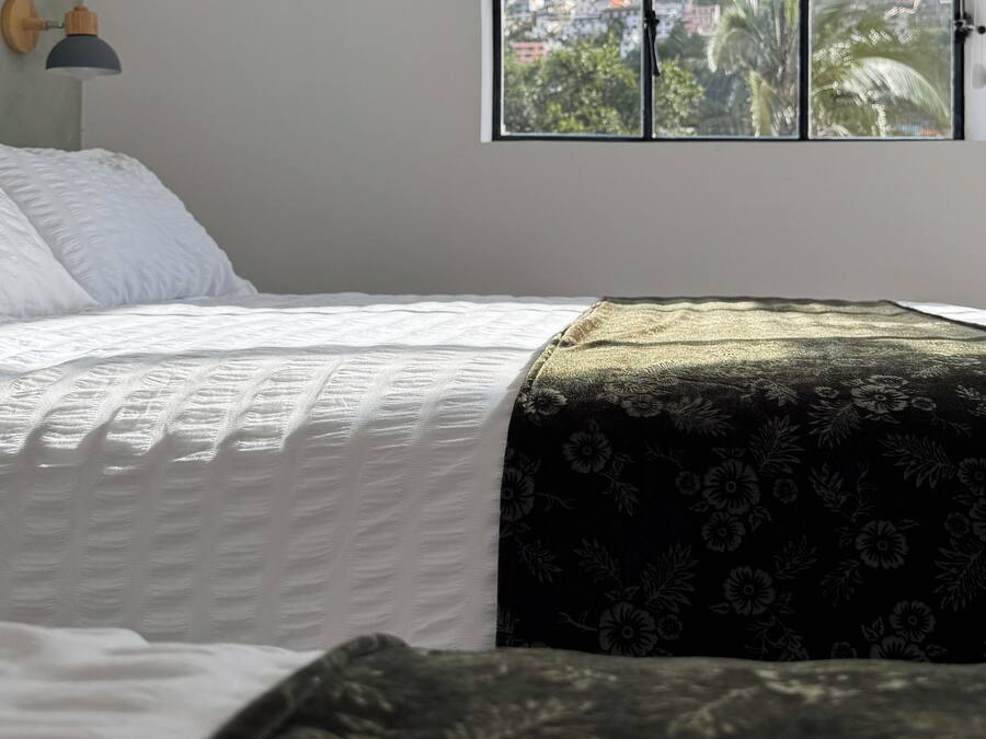 A bedroom with a bed, a window with a view of a statue and buildings, and a wall-mounted lamp.