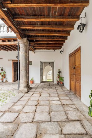 Une cour dallée de pierres, dotée d’un plafond en bois et de colonnes, qui mène à une arche ouverte.