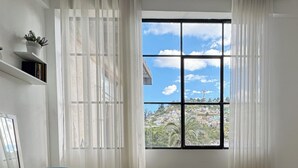 Quarto com uma mesa redonda e duas cadeiras azuis, janela com vista para a paisagem urbana e uma estante com uma planta.