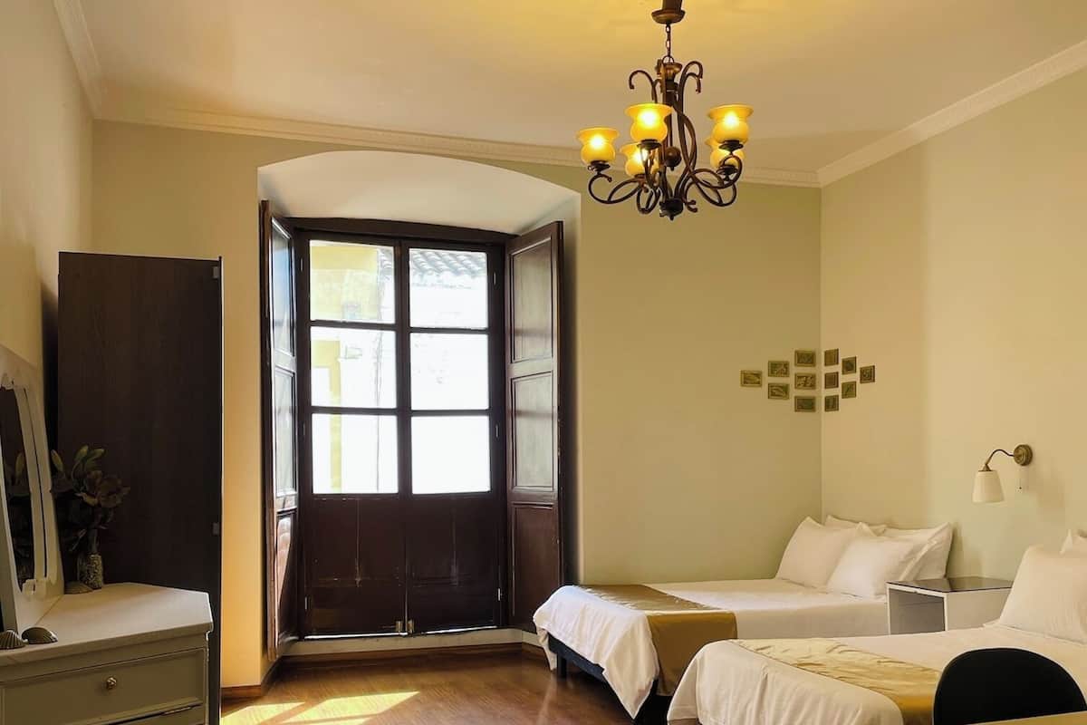A hotel room with a wooden floor, a chandelier, two beds, a desk, a dresser, and a large window with a view.