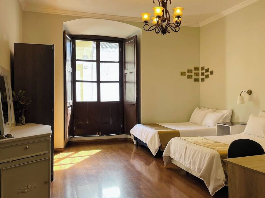 A hotel room with a wooden floor, a chandelier, two beds, a desk, a dresser, and a large window with a view.