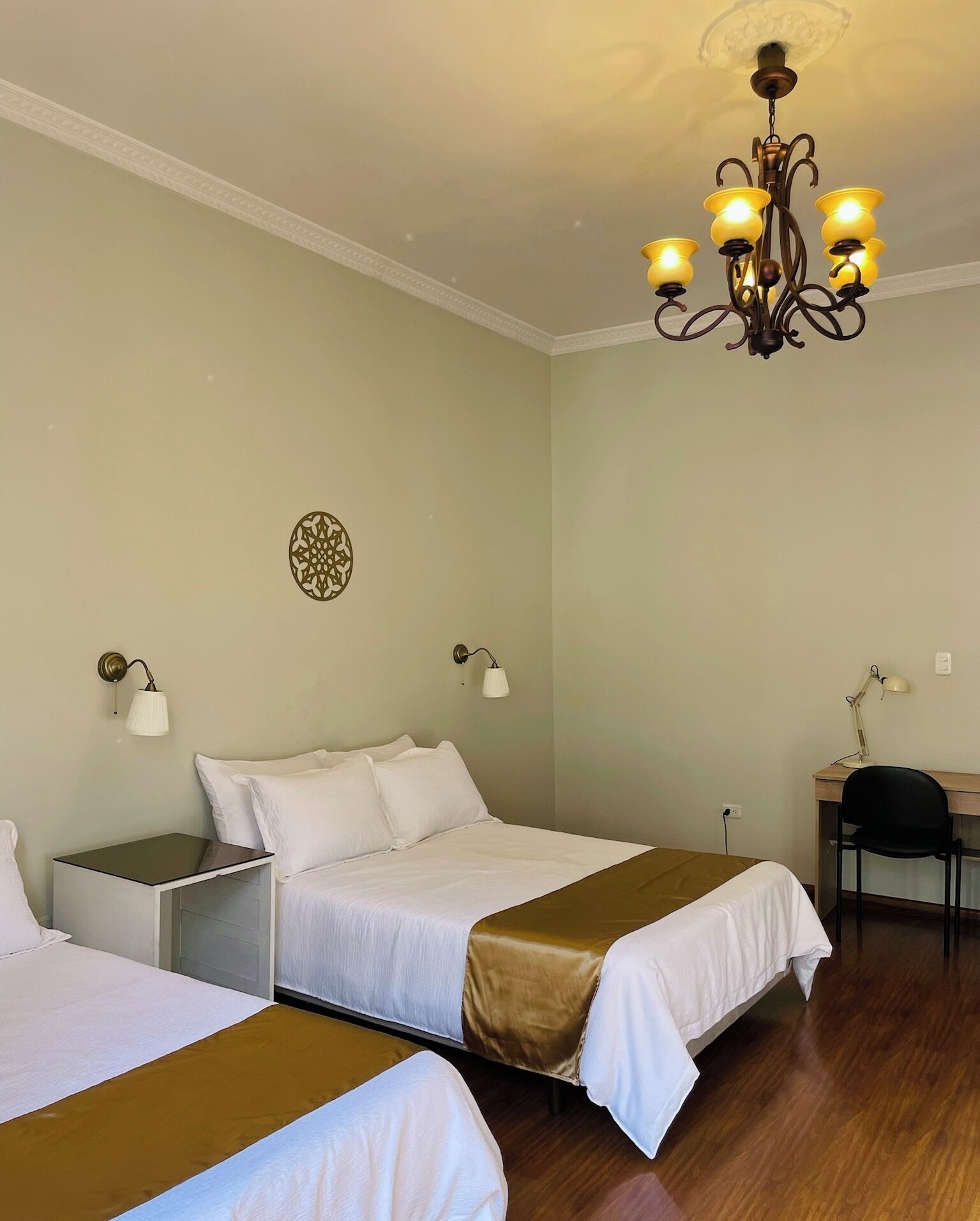 A hotel room with two beds, a desk, a chair, a chandelier, and a decorative wall piece.