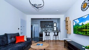 Una sala moderna con un sofá, una mesa de centro y un televisor. Se aprecia una cocina al fondo con un microondas y un lavamanos.
