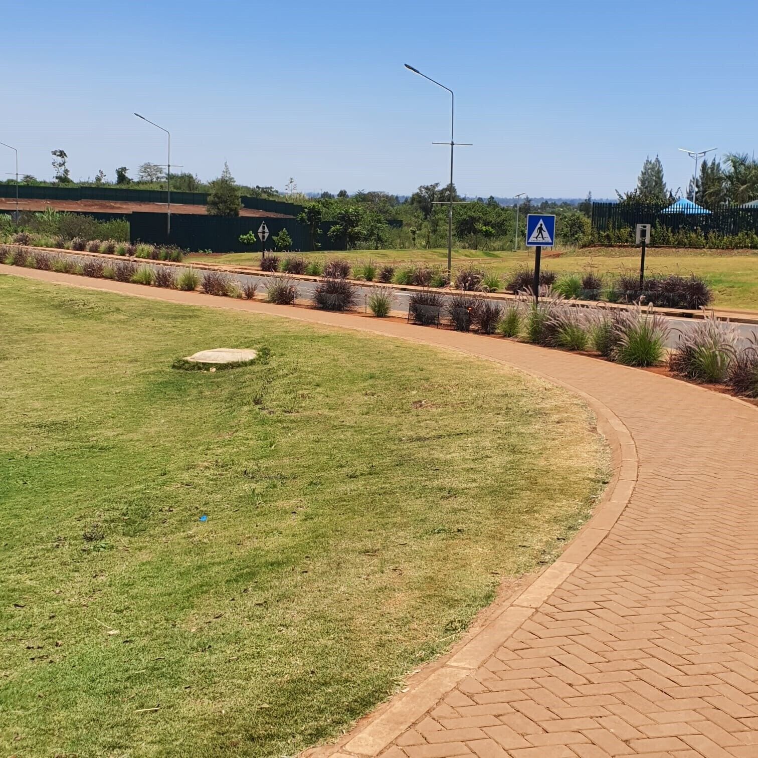 Uma trilha pavimentada serpenteando por uma área ajardinada com grama e plantas, um sinal de faixa de pedestres e postes de iluminação.