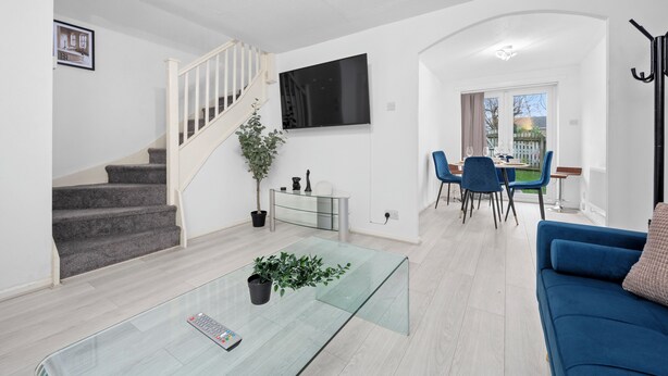 A modern living room with a glass coffee table, a blue sofa, a staircase, a dining area with chairs, and a TV mounted on the wall.