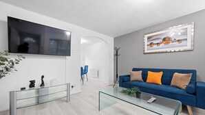 A modern living room with a blue sofa, glass coffee table, and a flat-screen TV mounted on the wall.