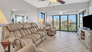 Een ruime woonkamer met een grote beigekleurige bank, een plafondventilator en een balkon met een tafel en stoelen.