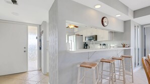 Een moderne keuken met witte kasten, een magnetron en een klok aan de muur. Er staan drie witte barkrukken met gevlochten zittingen.