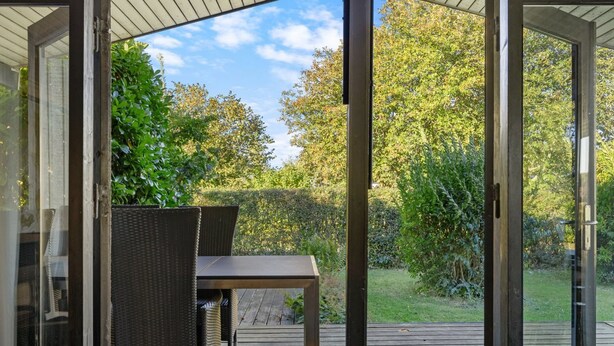 Een open terras met een tafel en stoelen, omgeven door glazen deuren en uitzicht op een tuin.
