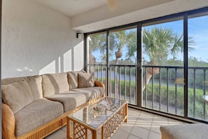 A balcony with a sofa and a glass coffee table, overlooking a body of water and palm trees.