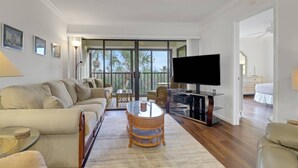 A living room with a sofa, a glass coffee table, a television on a stand, and a balcony with a view of palm trees. - 21400 Bay Village Dr 204 (Fort Myers Beach)