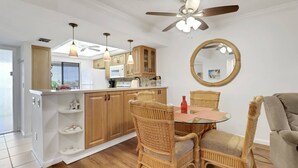 A compact kitchen with wooden cabinets, a dining table with wicker chairs, a round mirror, and a ceiling fan. - 21400 Bay Village Dr 204 (Fort Myers Beach)