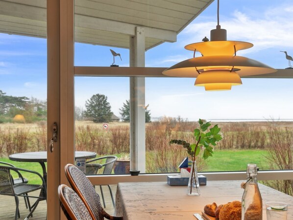 Een kamer met een groot raam dat uitkijkt op een veld, een tafel met stoelen, een hanglamp en een fles op de tafel.