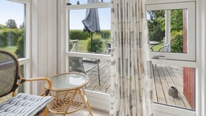 Une pièce avec une table en verre et des chaises en osier, de grandes fenêtres avec des rideaux, et une vue sur une terrasse en bois et un jardin.