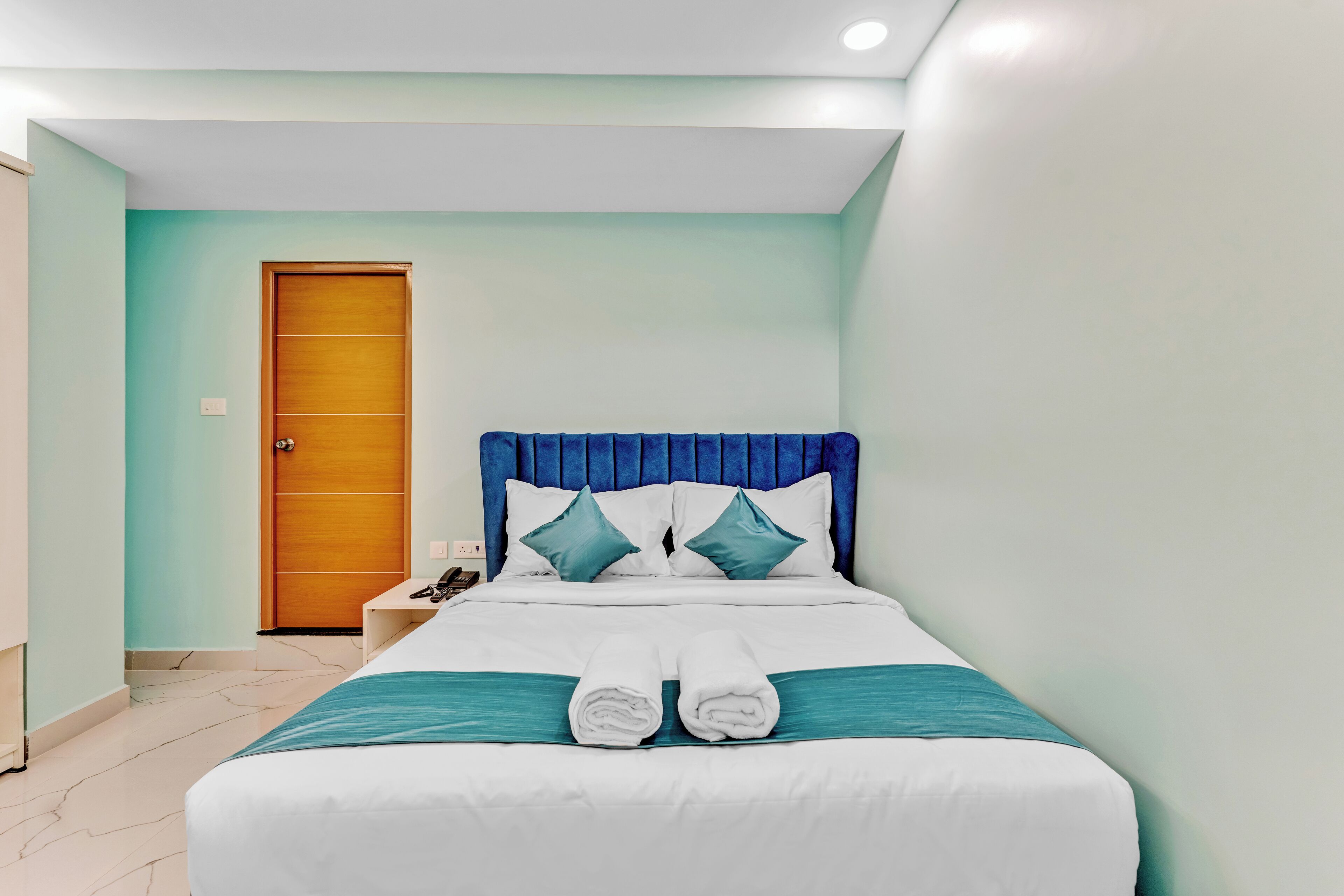 A neatly made bed with white linens and teal accents, a wooden door, and a nightstand with a phone.