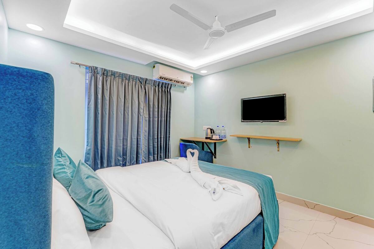 A hotel room with a bed, a ceiling fan, an air conditioning unit, a television, and a small table with a kettle and cups.