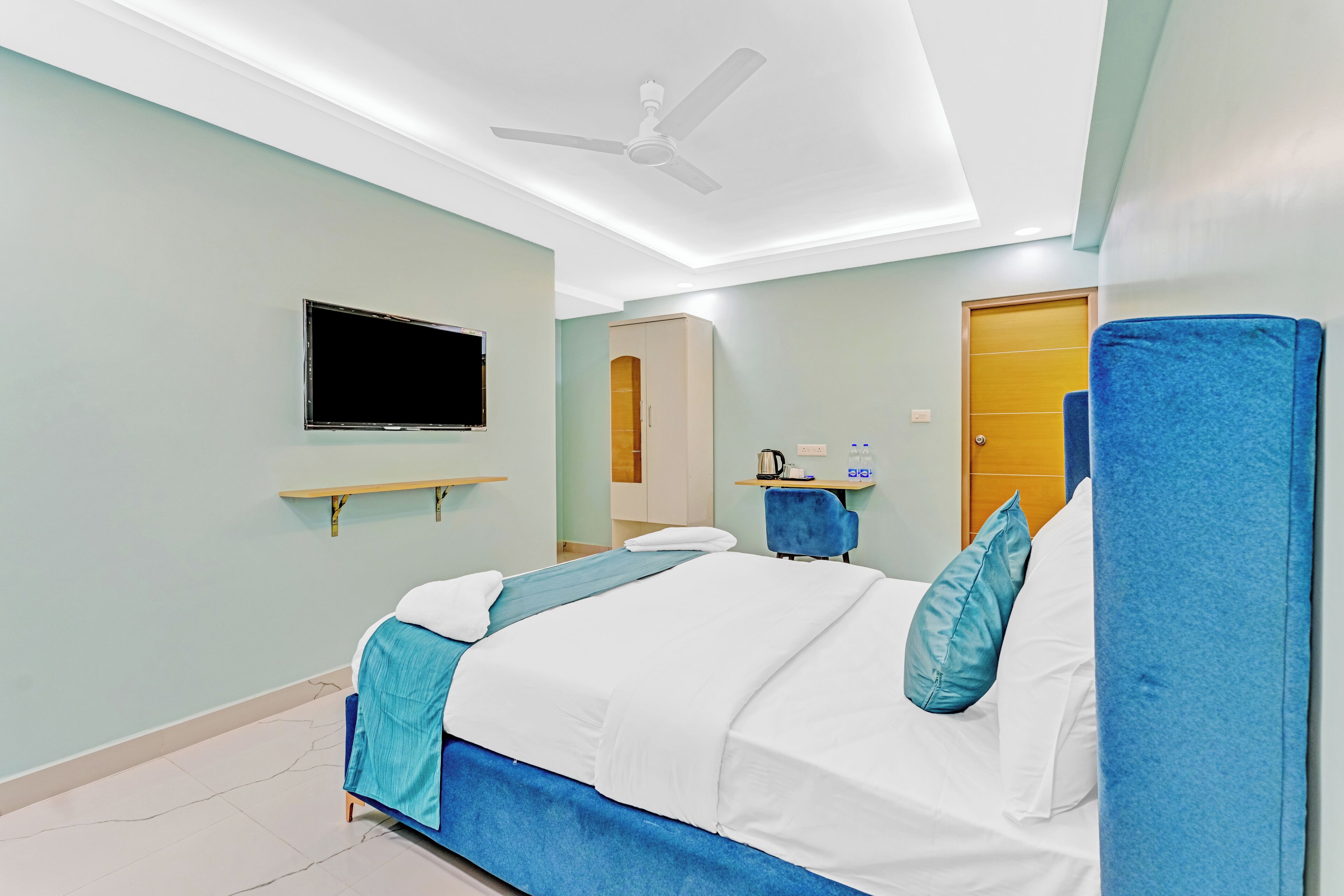 A hotel room with a bed, a television mounted on the wall, a ceiling fan, and a door.