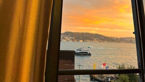A window with a view of a sunset over a body of water, a boat, and a distant shoreline. - Lokka Hotel (Istanbul)
