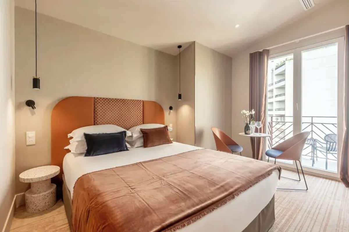 A modern hotel room with a large bed, a small round table, a chair, and a balcony with outdoor seating.