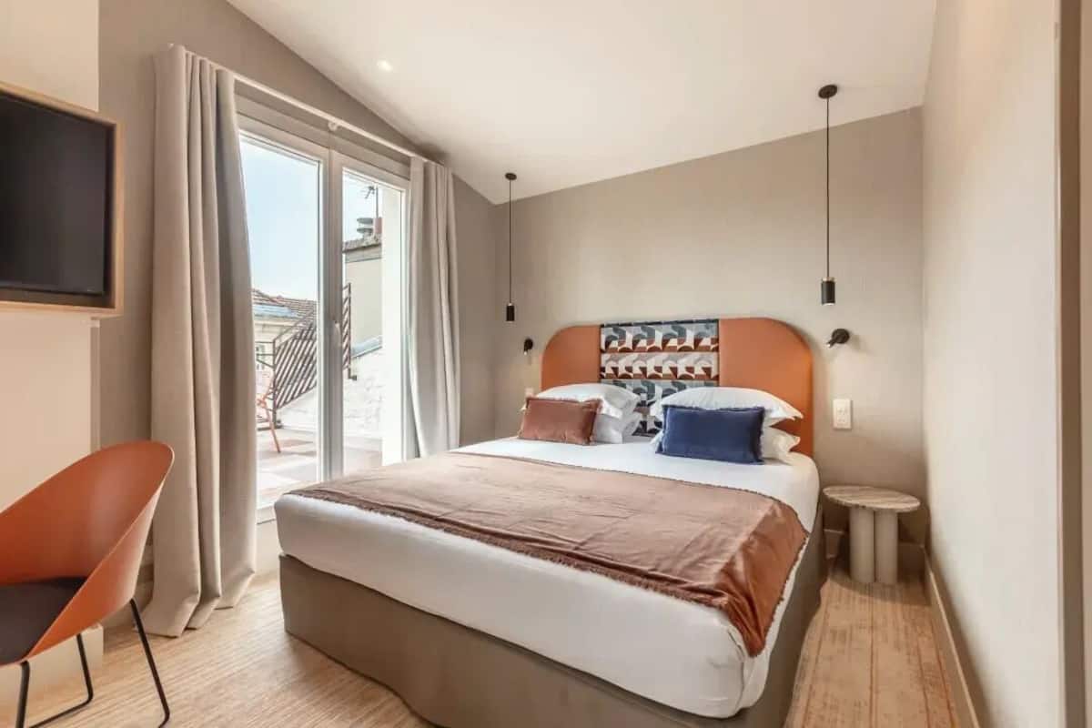 A hotel room with a bed, a chair, a small table, a TV, and a view of the outside.