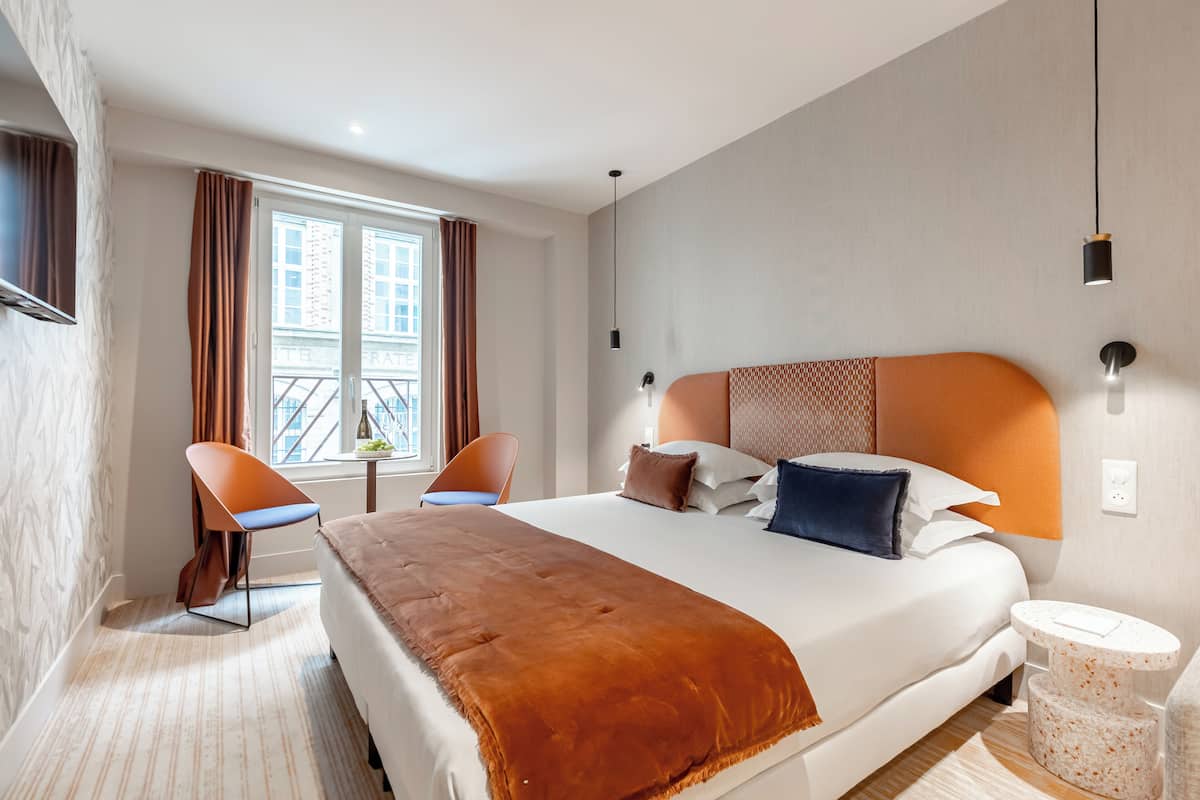 A hotel room with a large bed, a chair, a small table, and a window with curtains.