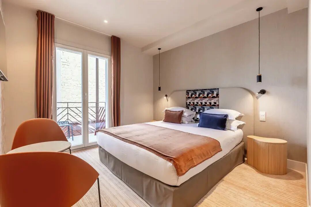 Una habitación de hotel moderna con una cama grande, una mesita pequeña y una silla. Tiene un balcón con cortinas y vistas al exterior.
