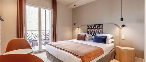Una habitación de hotel moderna con una cama grande, una mesita pequeña y una silla. Tiene un balcón con cortinas y vistas al exterior.