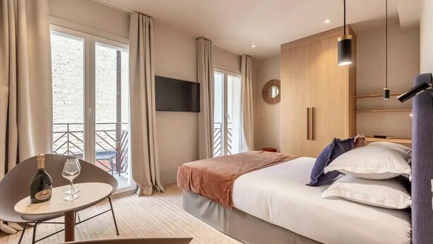 A modern hotel room with a bed, a TV, a balcony, and a table with a bottle and glass. - Suites FL by Sweett (Paris)
