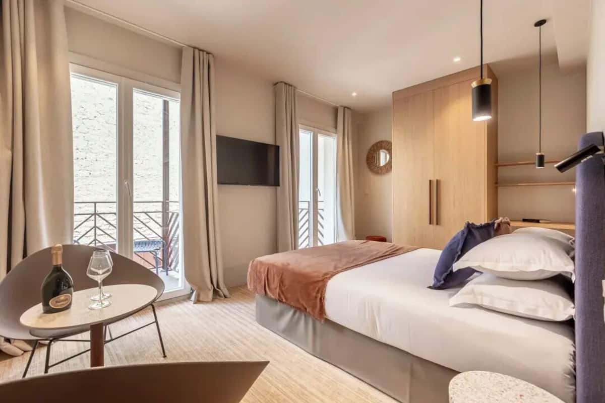 A modern hotel room with a bed, a TV, a balcony, and a table with a bottle and glass.