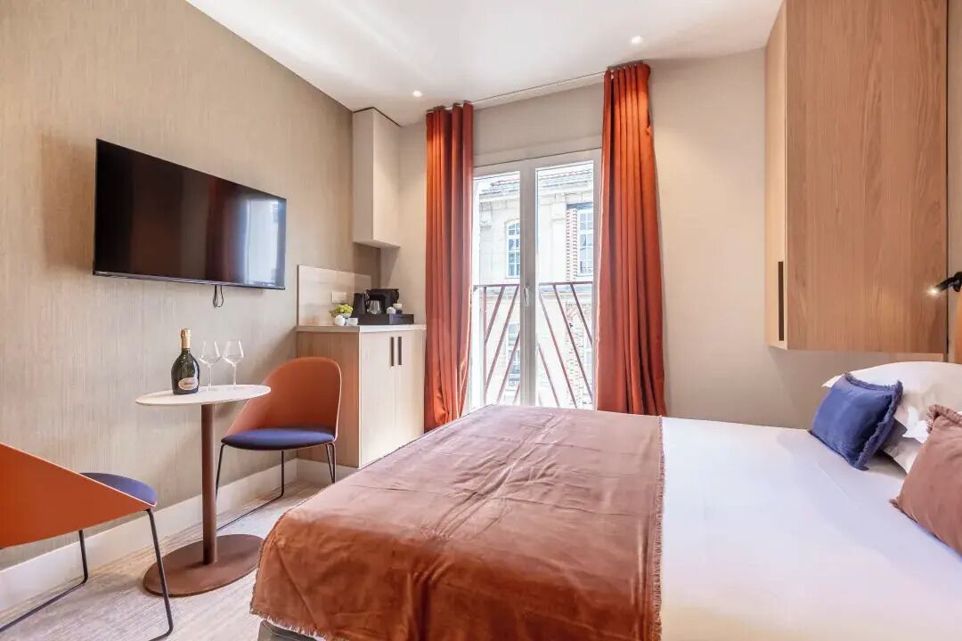 Habitación de hotel con cama, televisor montado en la pared, una mesita con una botella y vasos, una silla y un balcón con cortinas.