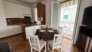 A dining area with a round table set for six, white chairs, a kitchen with white cabinets, and a sliding glass door to an outdoor area. - [200m from the Sea] 3 min from Porto Sole (Sanremo)