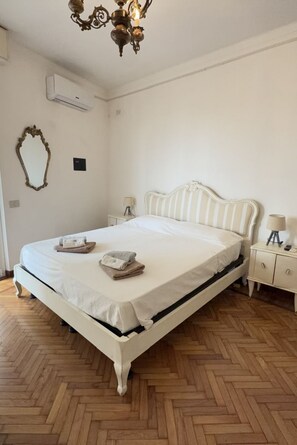 A bedroom with a white bed, a nightstand, a lamp, and a mirror on the wall.