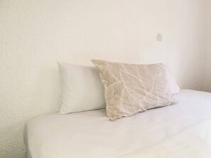 Una cama con dos almohadas, una lisa y otra con estampado de hojas, frente a una pared lisa.