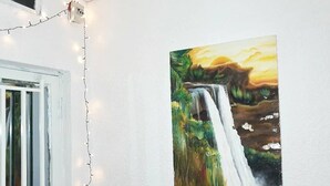 Una habitación con pared blanca, una ventana con persianas blancas y un cuadro de una cascada.