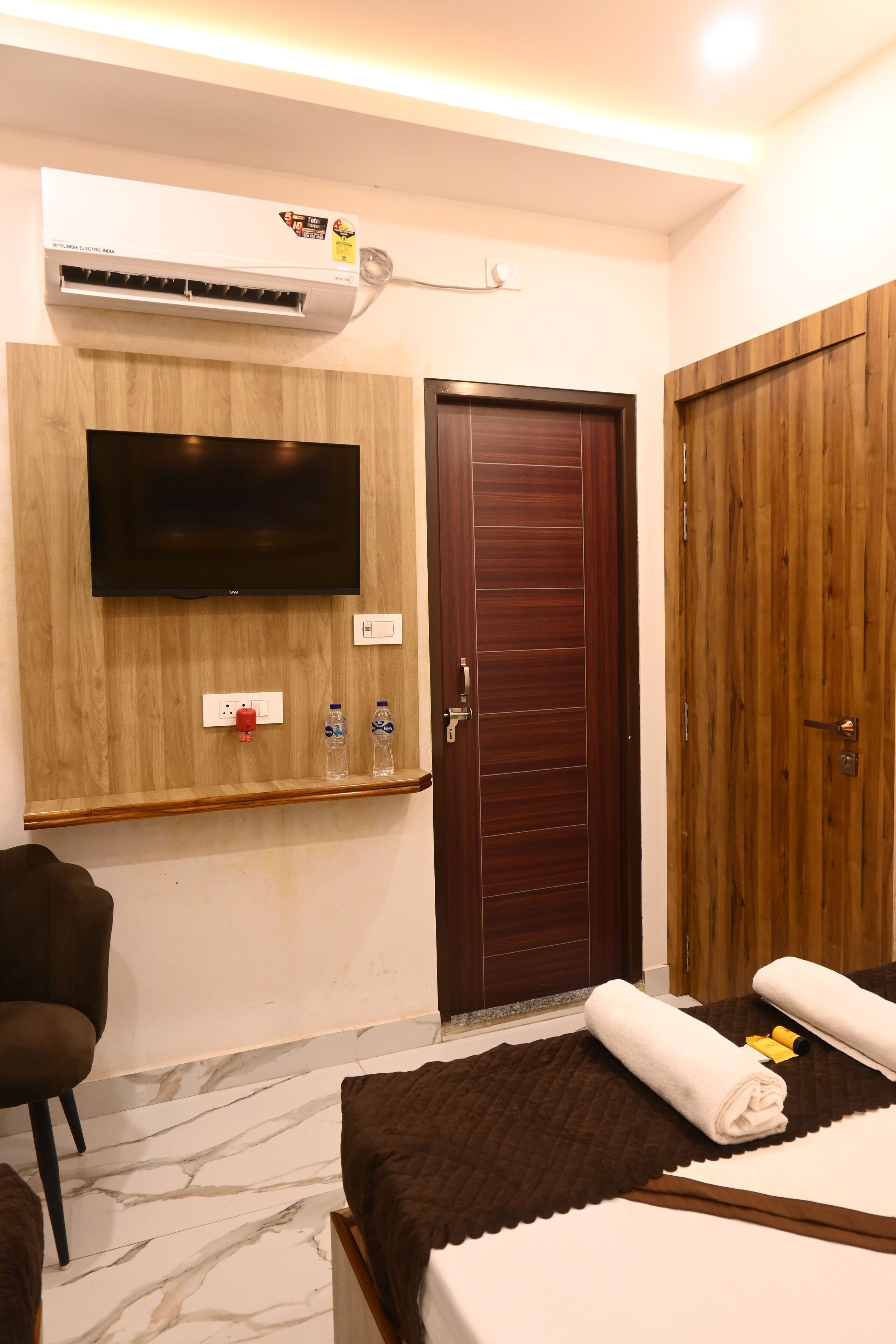A hotel room with a TV mounted on the wall, a wooden wardrobe, a bed with a brown bedspread, and a chair.