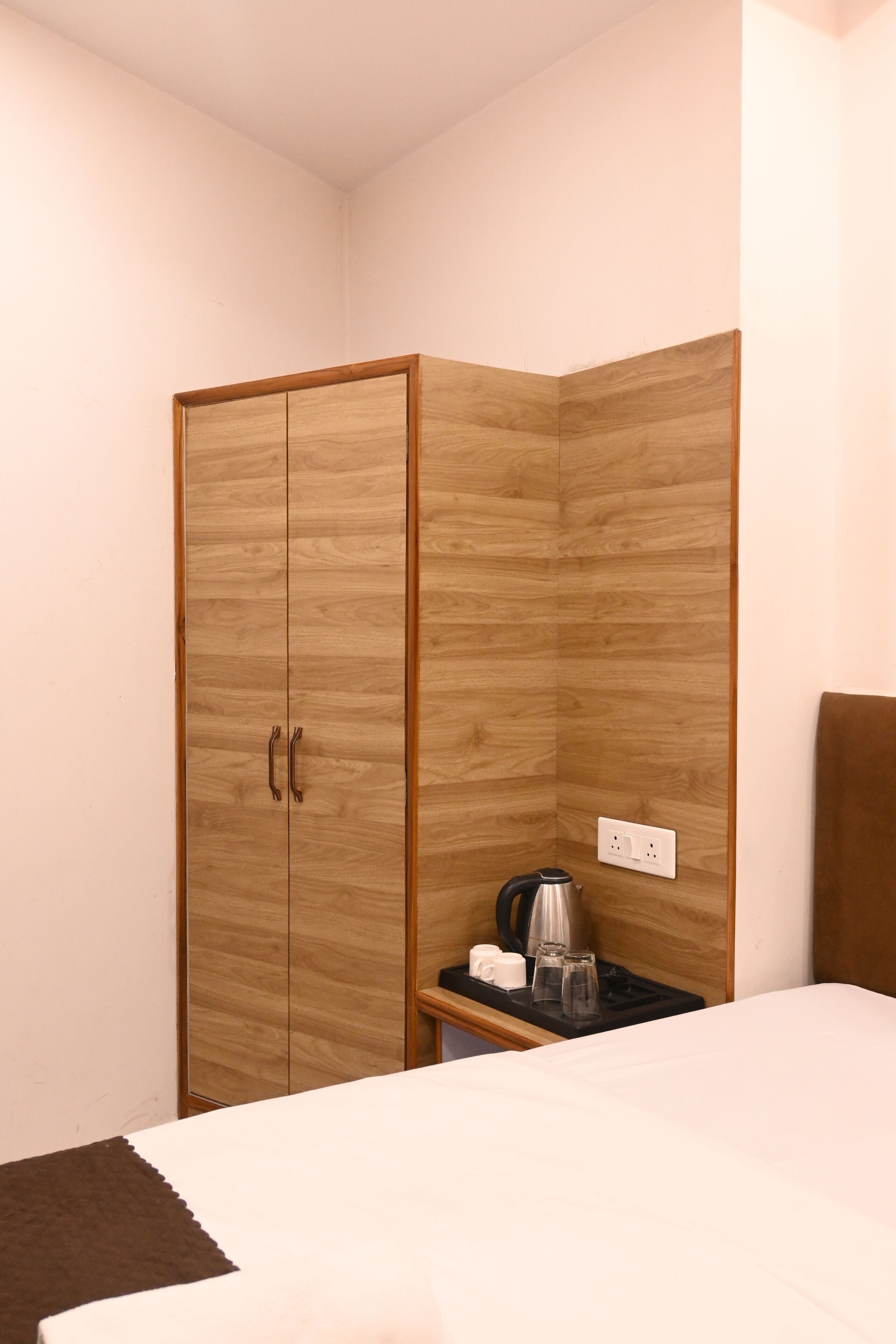 A bedroom with a wooden wardrobe, a bedside table with a kettle, and a power outlet.