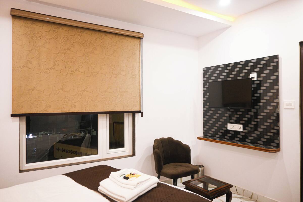 Ein Hotelzimmer mit großem Fenster, das mit einem beigen Rollo verdeckt ist, einem braunen Sessel, einem kleinen Holztisch und einem an der Wand befestigten Fernseher.