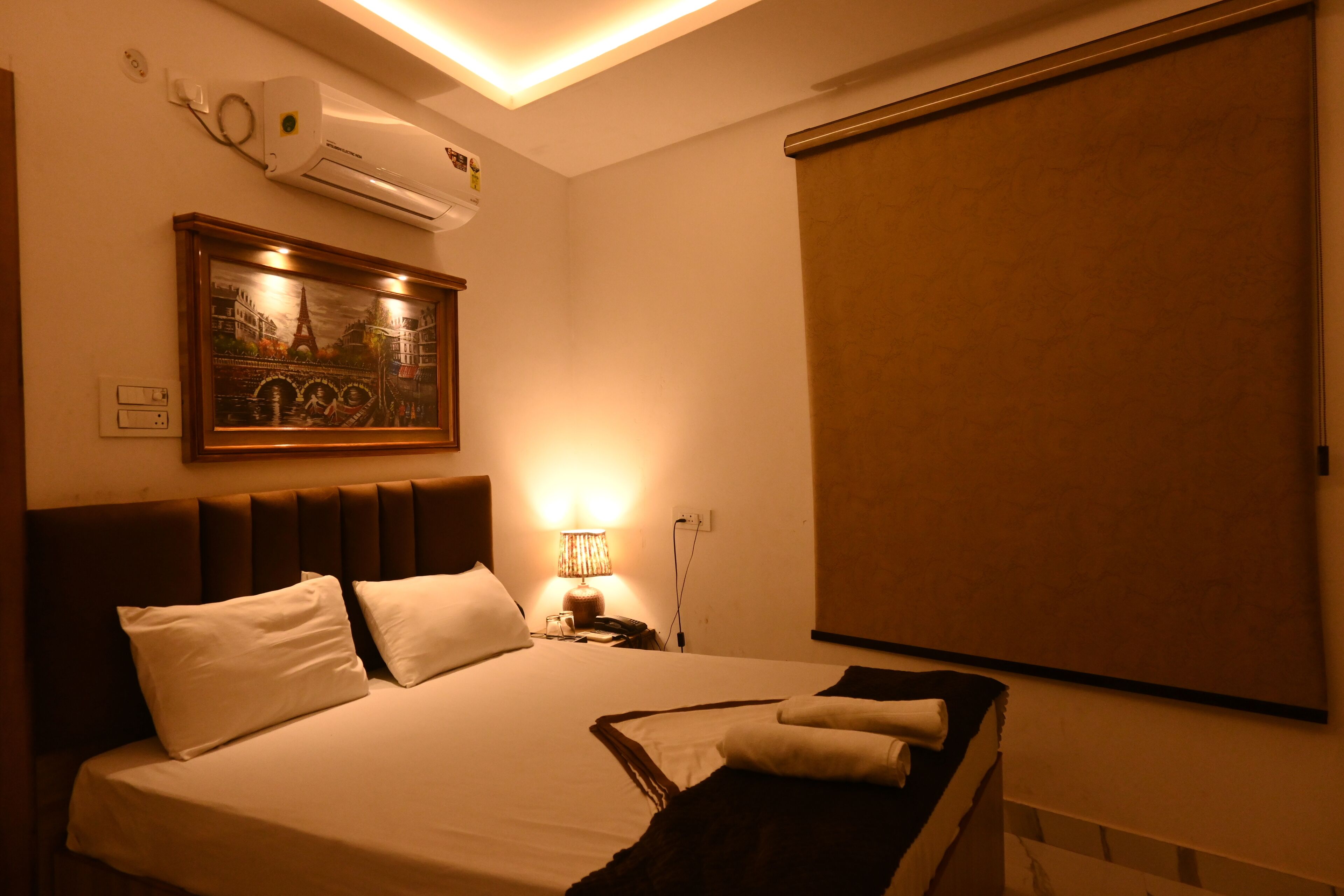 Ein Hotelzimmer mit einem Bett, einem Nachttisch, einer Lampe, einer Klimaanlage und einem großen Fenster mit Jalousie.